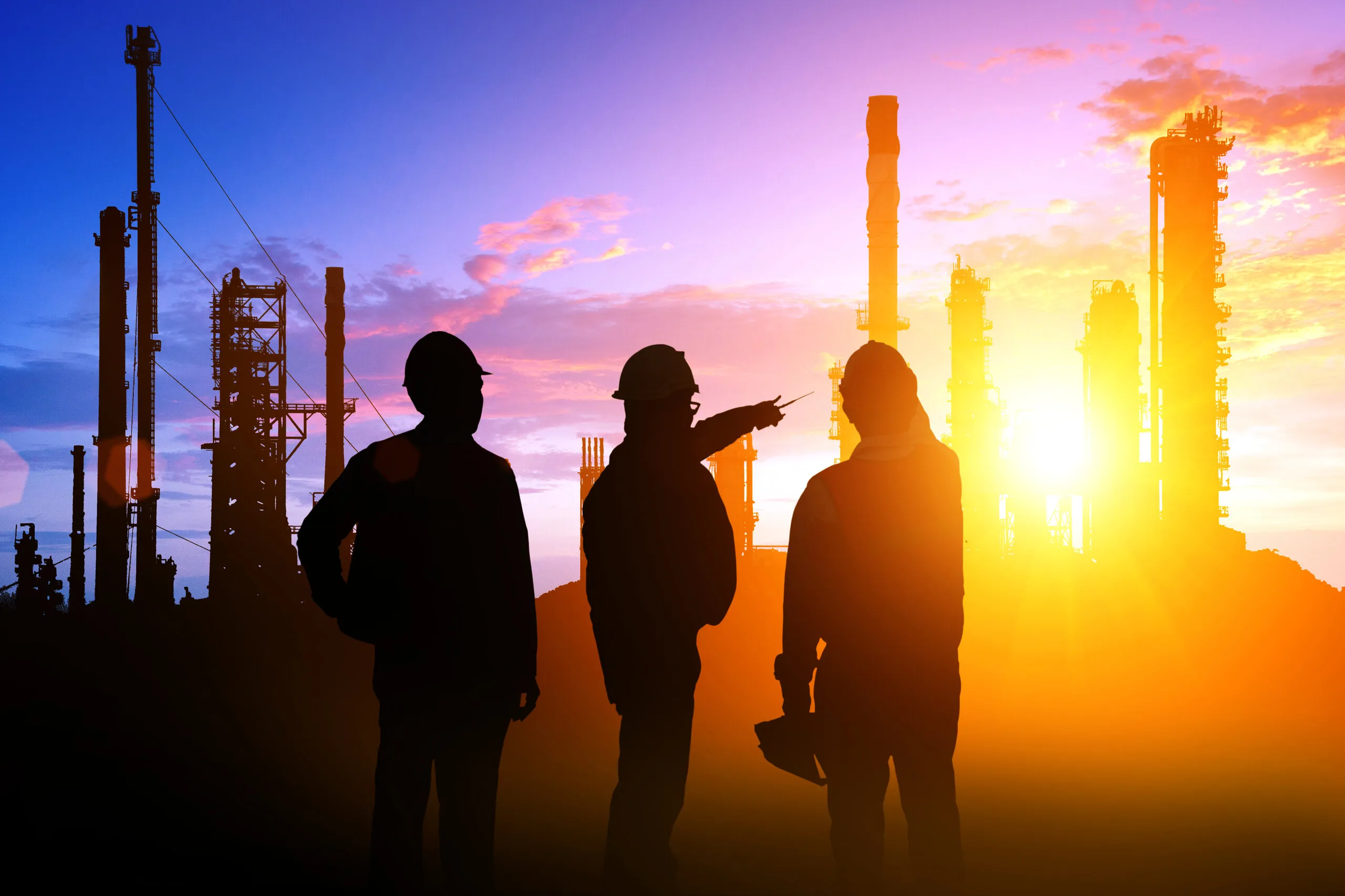 Three workers in hard hats silhouetted against a sunset at an industrial plant